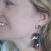 A close-up shot of a person wearing custom-made plague doctor mask earrings. The dangling earrings feature the classic long-beaked doctor mask, highlighting the detailed crafting.