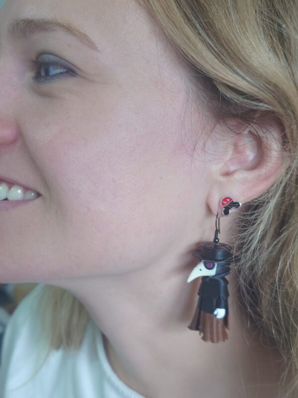 A close-up shot of a person wearing custom-made plague doctor mask earrings. The dangling earrings feature the classic long-beaked doctor mask, highlighting the detailed crafting.